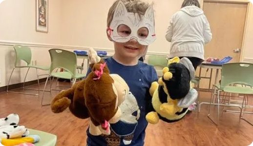 A kid in a paper mask plays with puppets in a classroom.